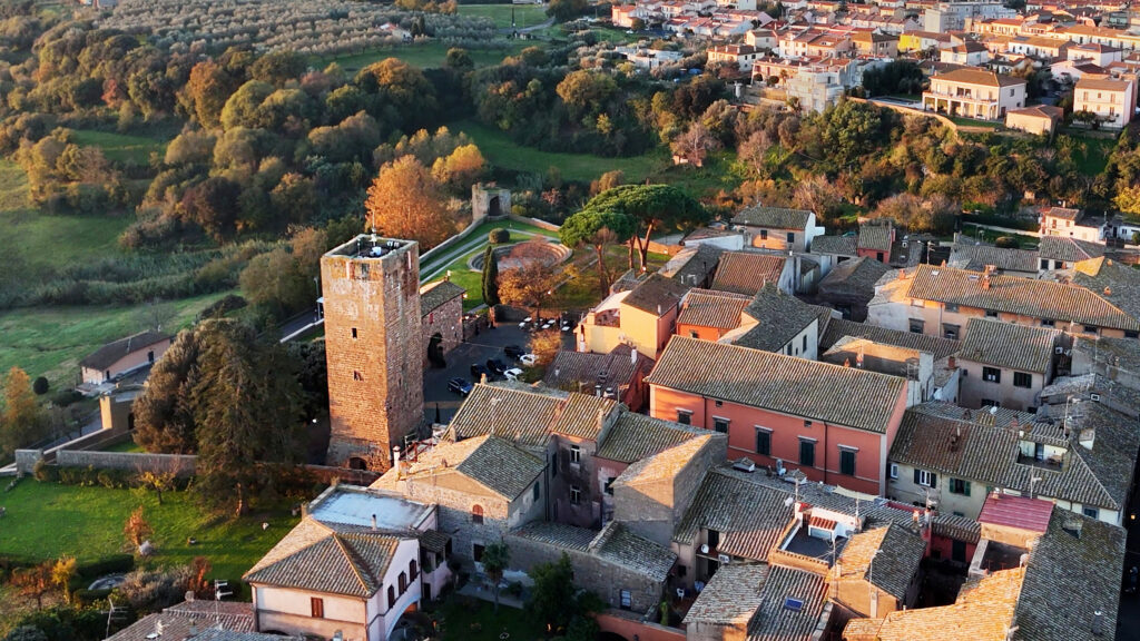 Ripresa con drone della Torre di Lavello e dei tetti circostanti nel centro storico di Tuscania, che risplendono nella luce dorata della sera, con le verdi colline sullo sfondo.