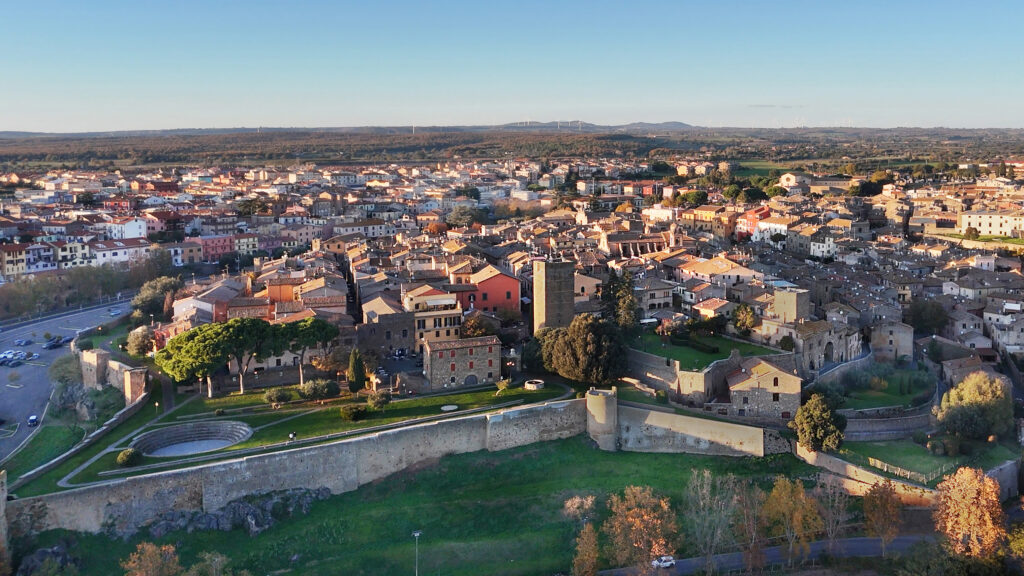 Veduta aerea del centro storico di Tuscania con la Torre di Lavello, le mura medievali e l'anfiteatro nella calda luce della sera, circondato da dolci colline e tetti.