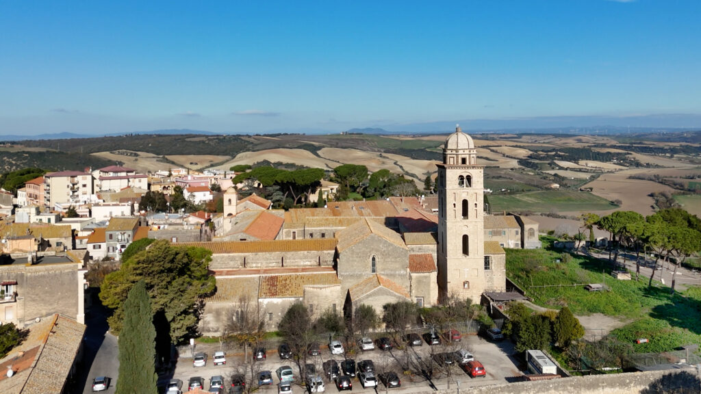 Drohnenaufnahme der Kathedrale Santa Margherita in Tarquinia mit ihrem markanten Glockenturm, umgeben von der hügeligen Landschaft Latiums und einem Parkplatz im Vordergrund.