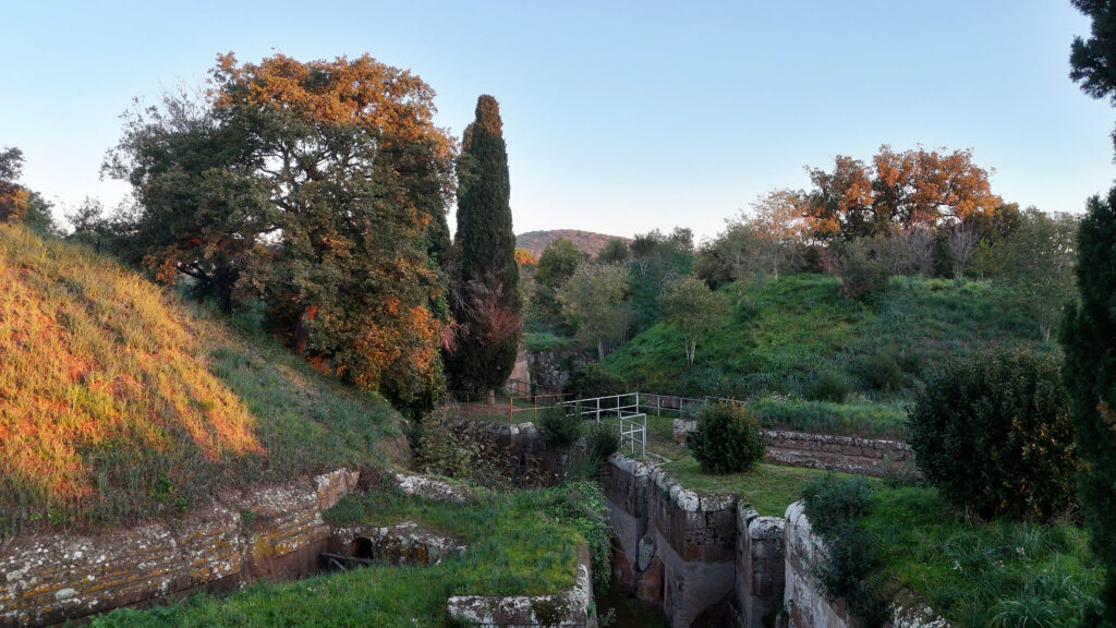 Blick auf die etruskische Nekropole von Cerveteri bei Abendlicht: Antike Gräber zwischen herbstlich gefärbten Bäumen, grasbedeckten Hügeln und einem klaren Himmel im Hintergrund.