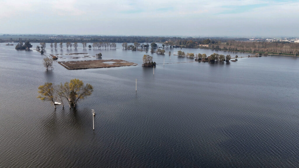 Aerial view of flooded farmlands in the Po Delta with scattered trees, a small boat, and submerged power lines, set against the flat horizon of Emilia-Romagna.