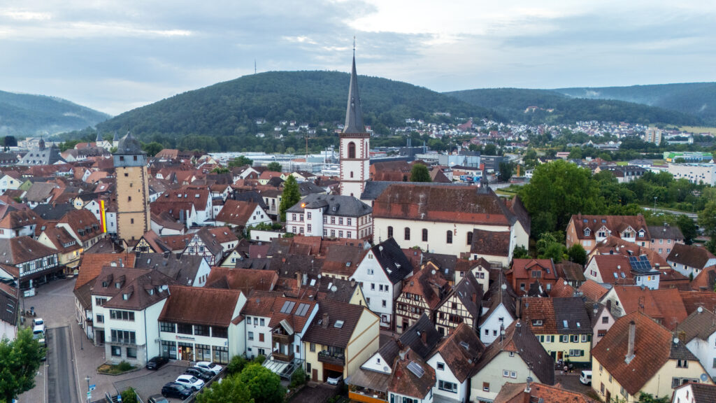 Vista da drone del centro storico di Lohr am Main con la chiesa di San Michele e la torre medievale, circondata da tetti rossi e da lontane colline verdi.