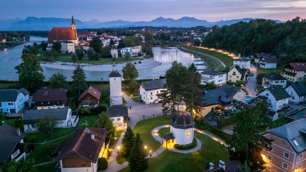 View of the Silent Night Chapel in Oberndorf, Salzburg, Austria, surrounded by lights and the Salzach River at blue hour.