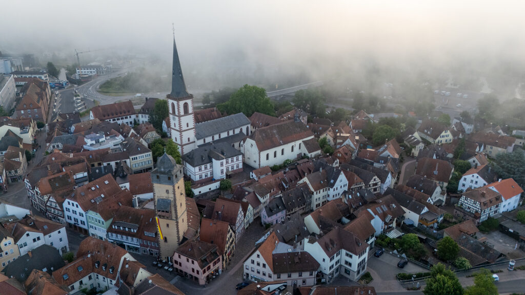 A quiet aerial view of a half-timbered town on the Main, showing the historic old town of Lohr am Main as church towers and half-timbered houses emerge from the morning fog in Lower Franconia.