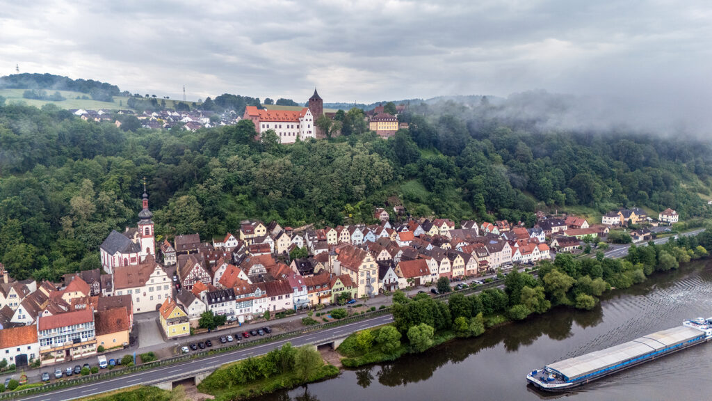 Aerial view of Rothenfels with historic houses along the Main river, Burg Rothenfels above the town and fog drifting through the forested hillside